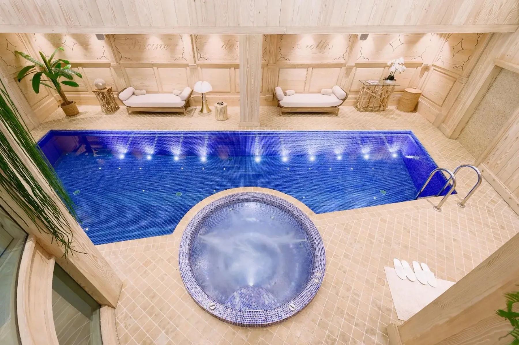 Luxurious indoor spa with a blue-lit pool, a round jacuzzi, loungers, and elegant wooden decor.