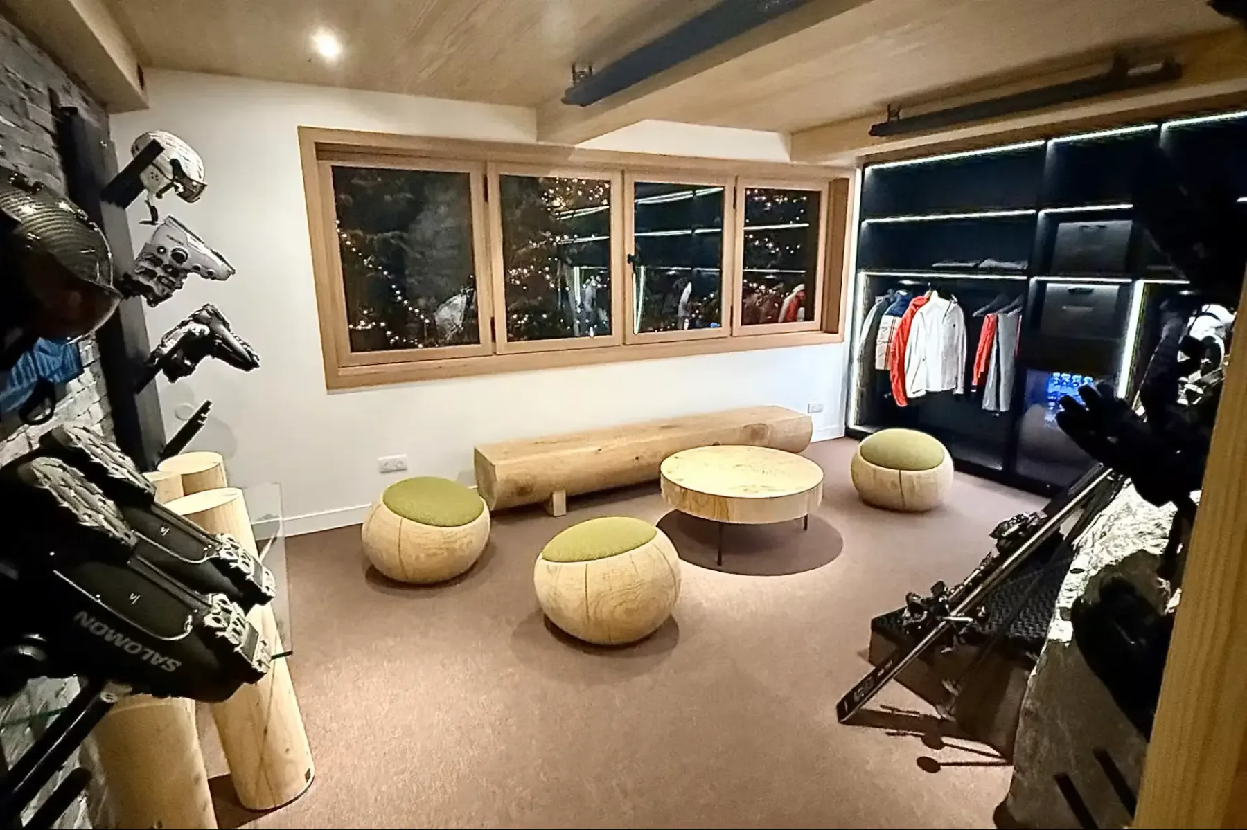 A cozy ski lodge room with wooden stools, a log bench, round tables, and ski gear displayed on racks, with large windows and soft lighting.