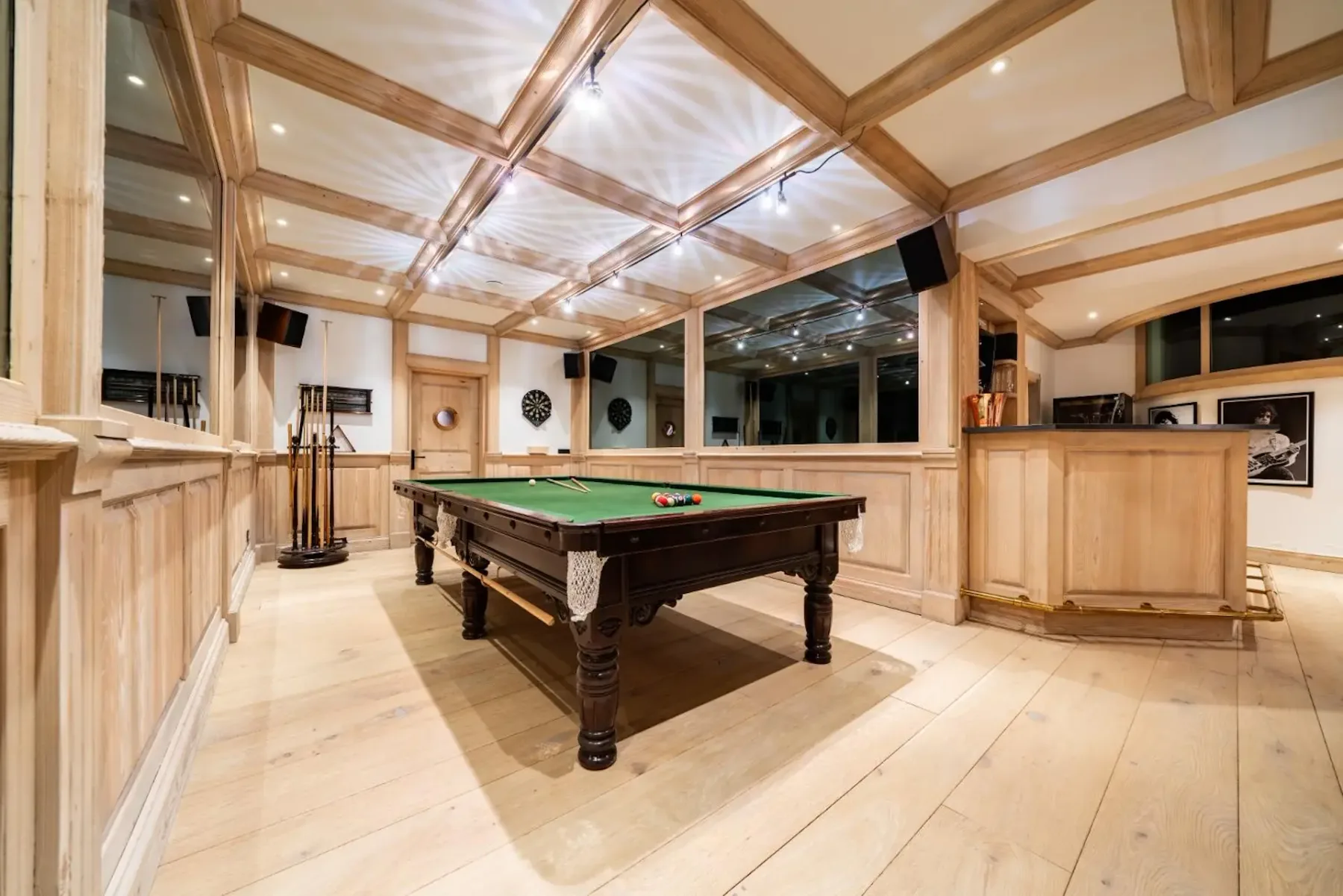 A spacious game room with a billiards table, wooden paneling, dartboard, and ceiling lights on a polished wooden floor.