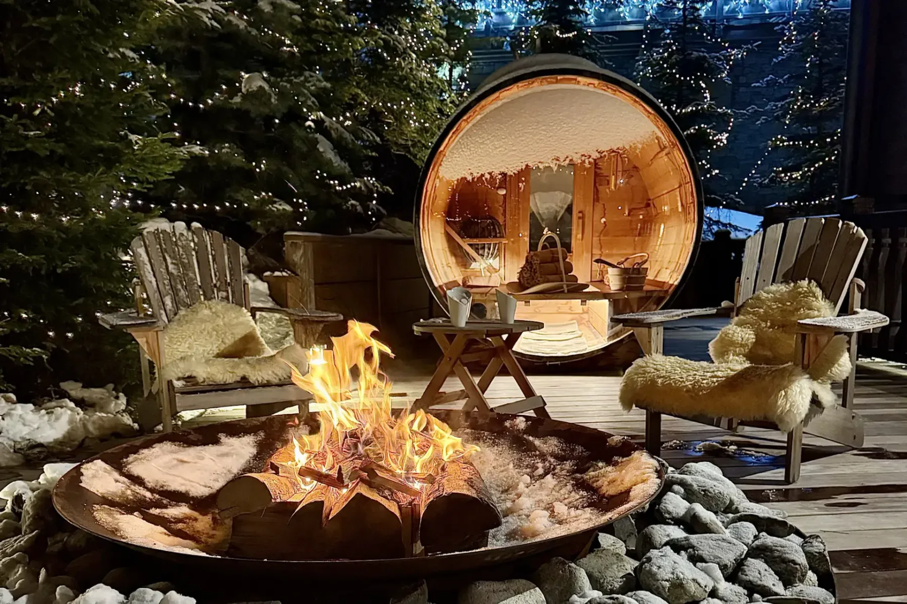 Cozy outdoor scene with a fire pit, wooden chairs with fur throws, and a barrel sauna, surrounded by snow and string lights.