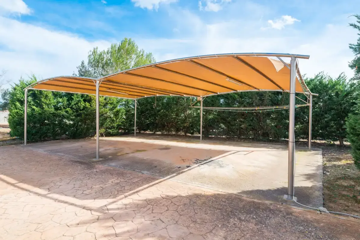 An outdoor paved patio sits beneath a broad orange canopy, with greenery behind it.
