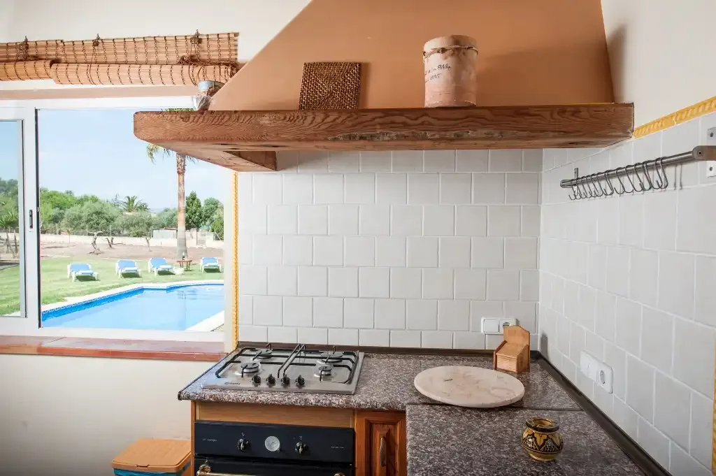 A kitchen with a stovetop beneath a wooden shelf and a window overlooking a pool.