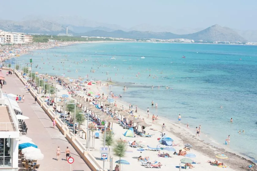 A crowded beach stretches along a turquoise bay with mountains in the distance.