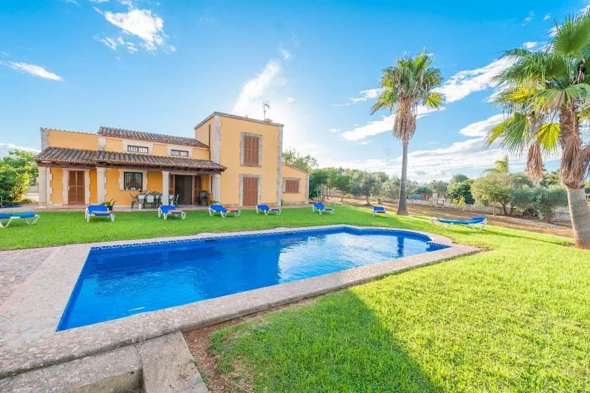 A swimming pool sits in a grassy yard beside a large tan house.