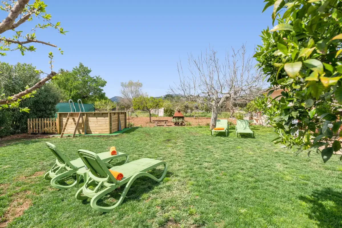 Un jardín con césped, tumbonas verdes, árboles y una pequeña estructura de madera para jugar.