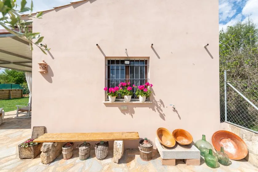 Un patio soleado con una ventana pequeña, jardineras con flores y asientos de madera.