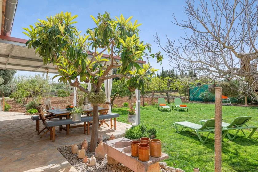 Un patio ajardinado con una mesa bajo los árboles y tumbonas sobre el césped.
