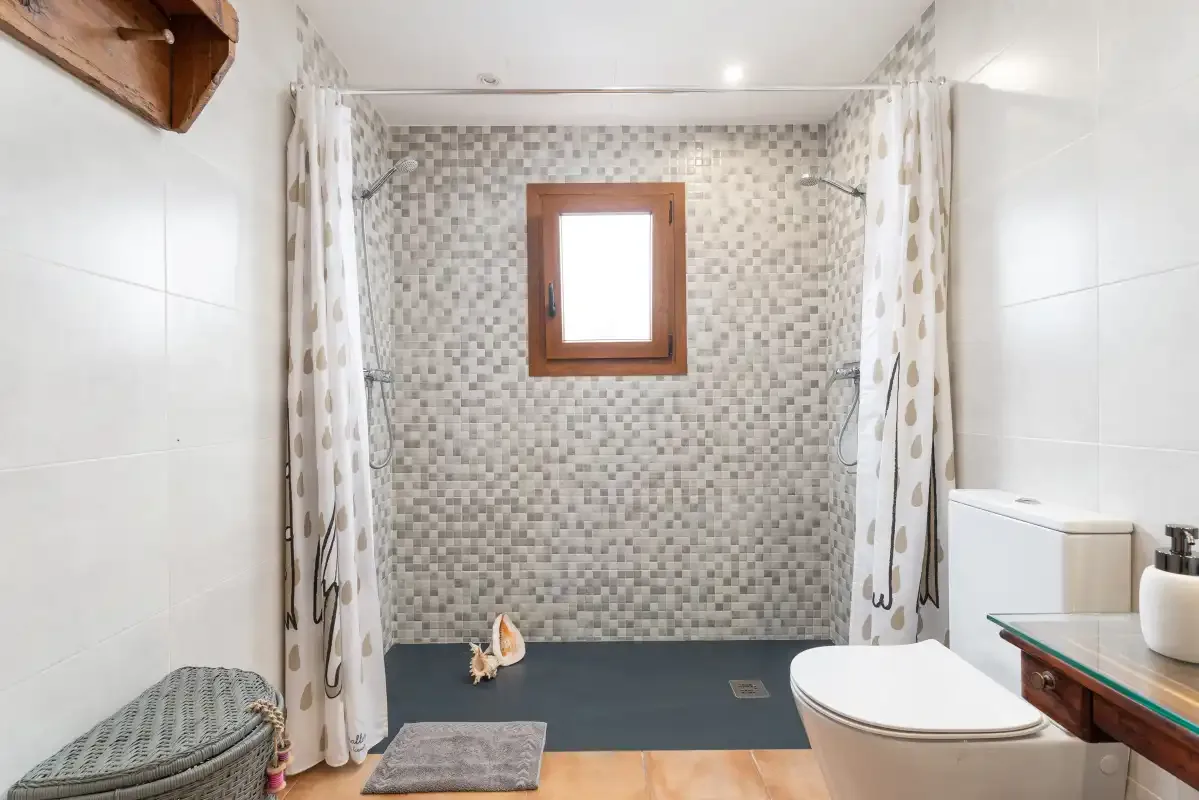 Un baño con una zona de ducha revestida de azulejos, inodoro y una ventana pequeña.