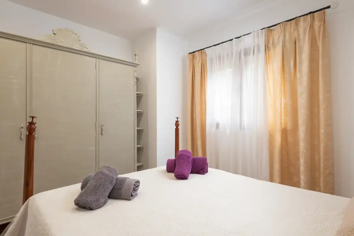 Dormitorio con una cama blanca, toallas dobladas y ventanas con cortinas.