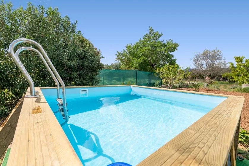 Una piscina al aire libre con una tarima de madera, vegetación y cielo azul.