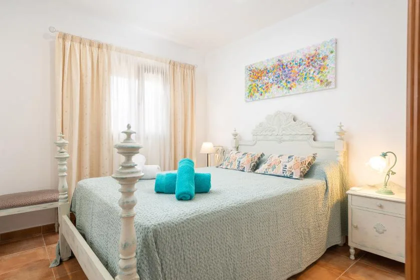 Un dormitorio luminoso con una cama bajo un cuadro colorido y una ventana con cortinas.