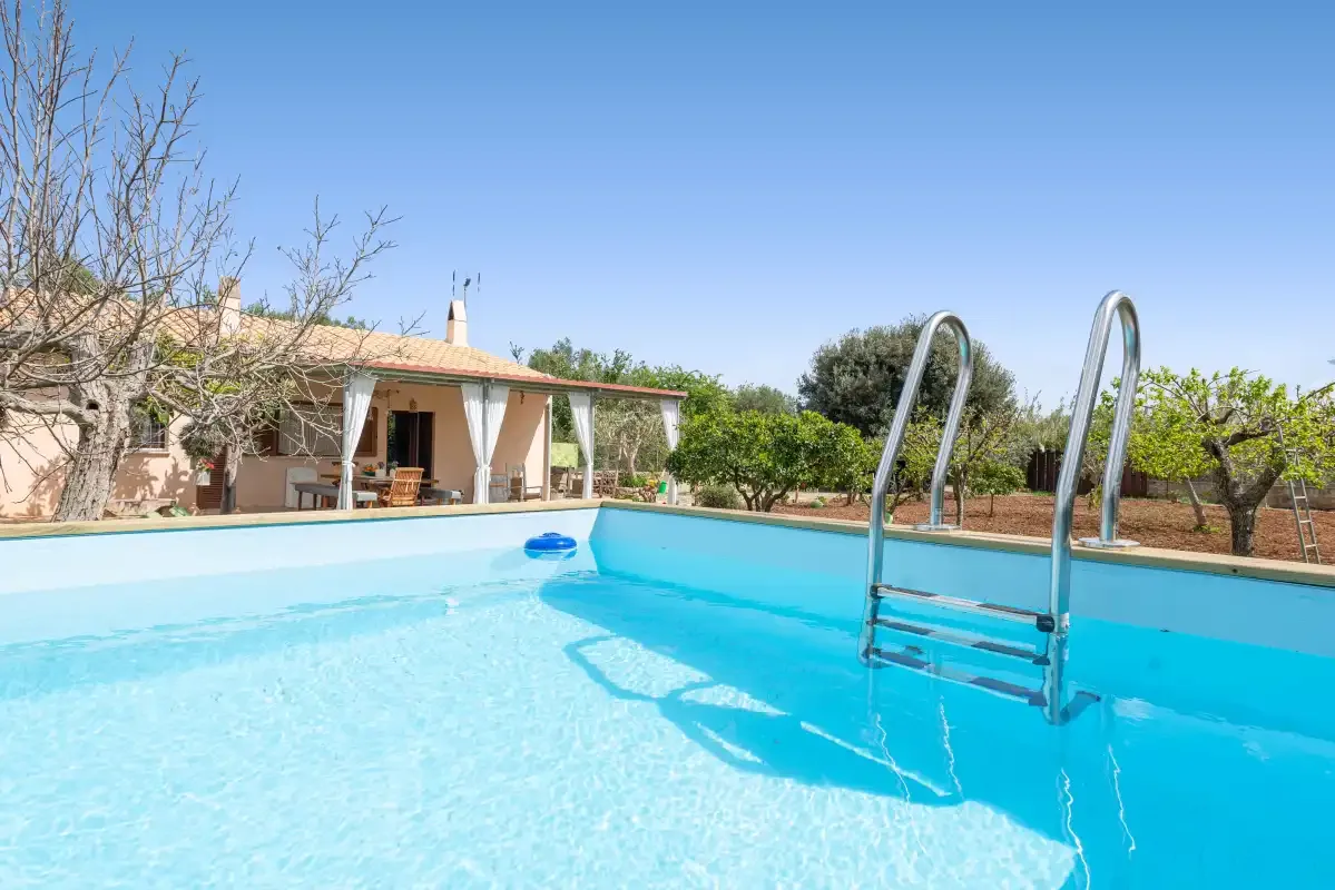 Una piscina ocupa el primer plano, con una casa pequeña y árboles al fondo.