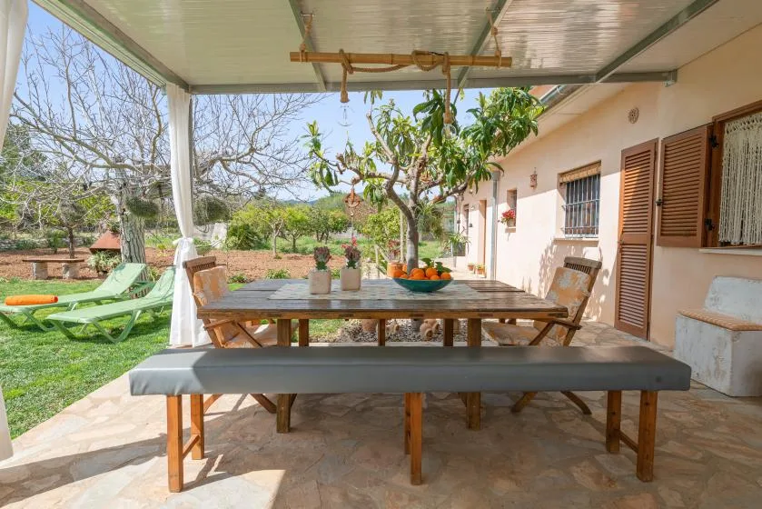 Una zona de comedor exterior cubierta, con mesa de madera y asientos, da a un jardín.