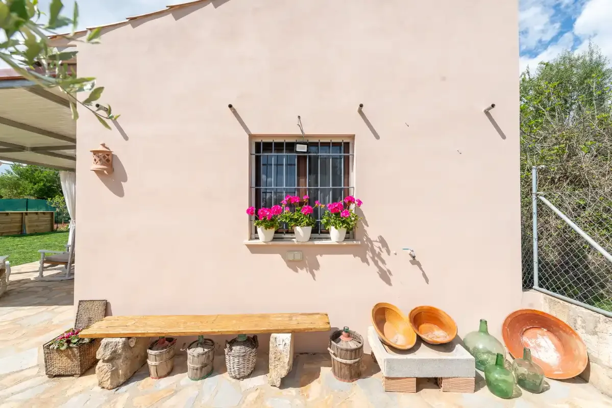 Una pared exterior clara tiene una pequeña ventana con barrotes y flores de colores.