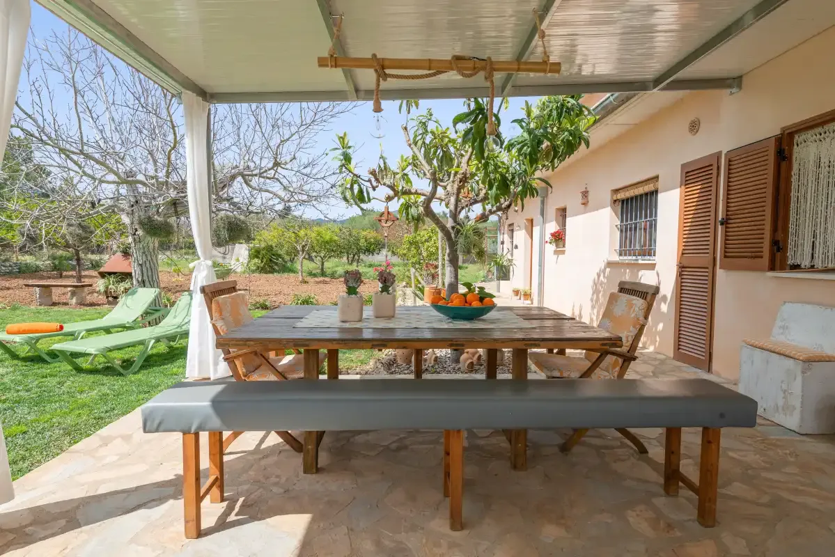 Un patio cubierto tiene una larga mesa de comedor de madera y césped verde al fondo.