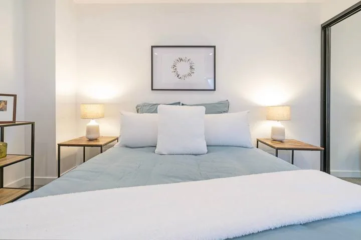 A neatly made bed sits between two bedside tables in a bright bedroom.