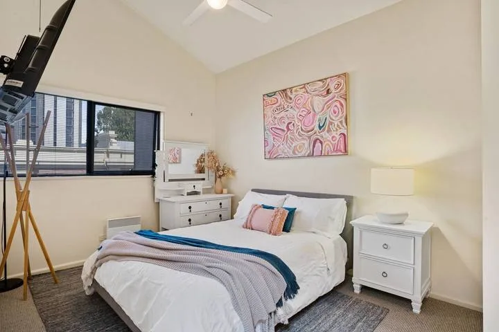 A bright bedroom with a bed, bedside table, and colorful artwork above.