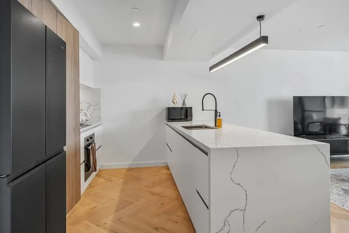 A modern kitchen with a white island, dark refrigerator, and light wood flooring.