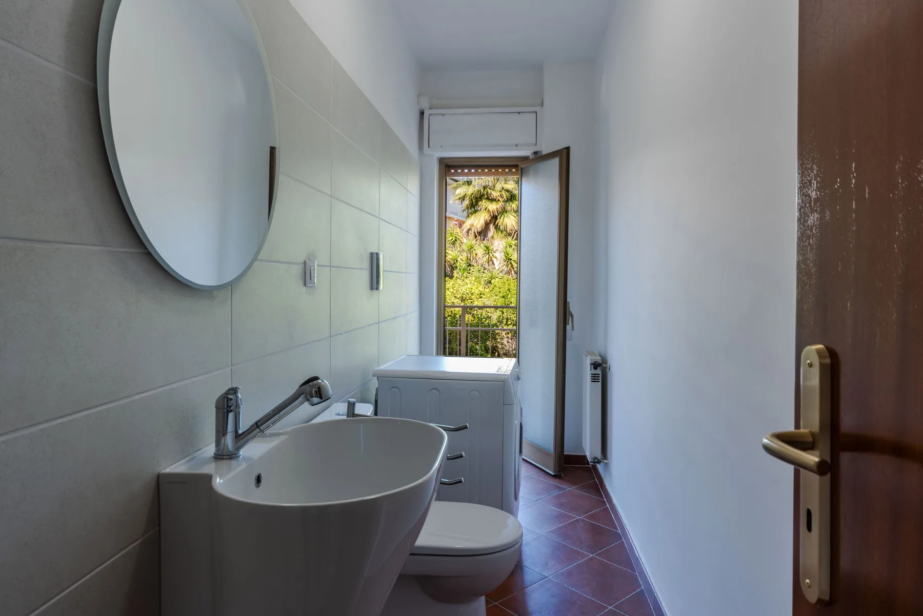 Un baño estrecho con lavabo e inodoro junto a una ventana abierta.