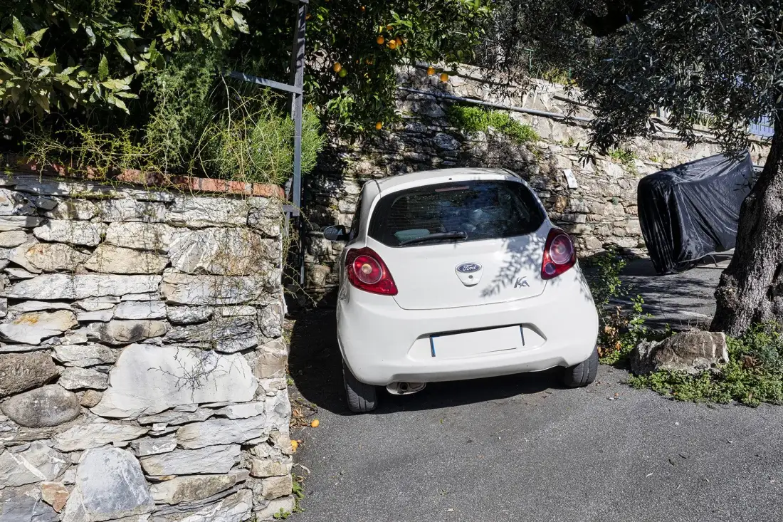 A white car is parked on a paved driveway beside a stone wall.