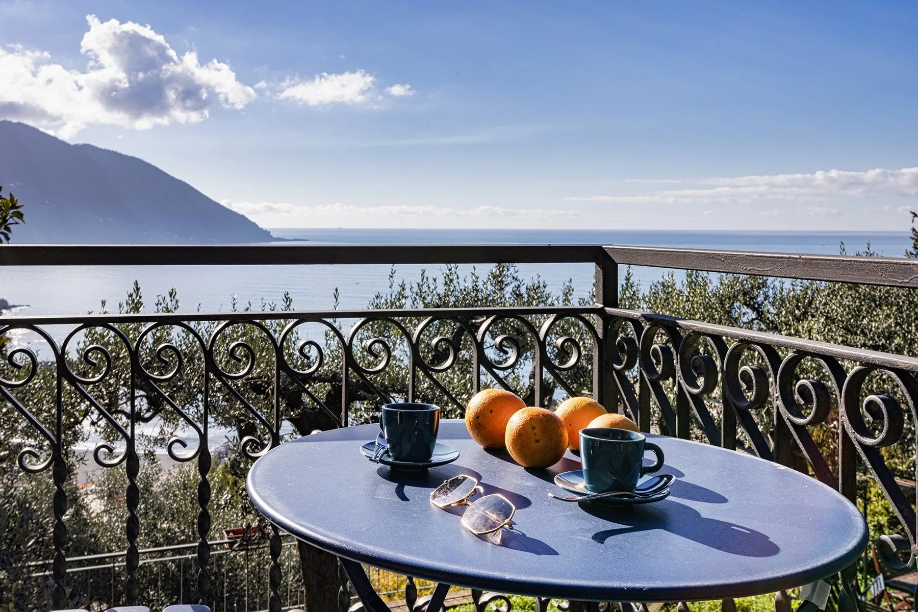 A balcony table with cups and fruit overlooks the sea and distant mountains.
