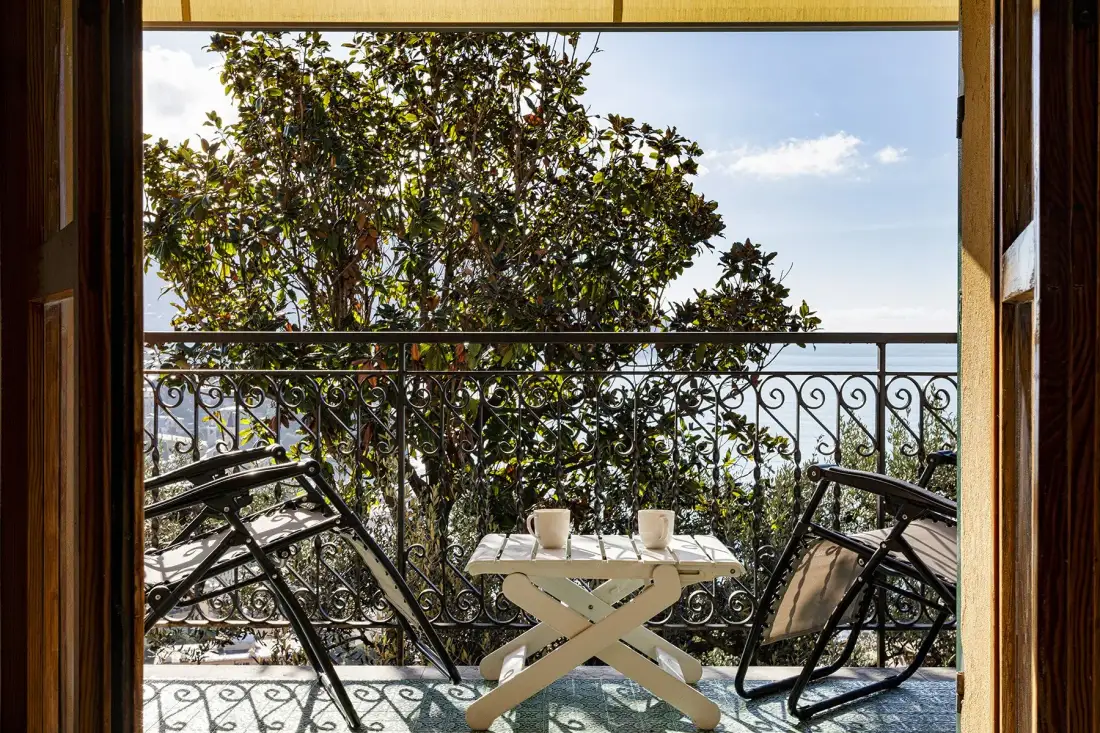 A small balcony with outdoor seating and a table overlooks leafy trees and the sky.