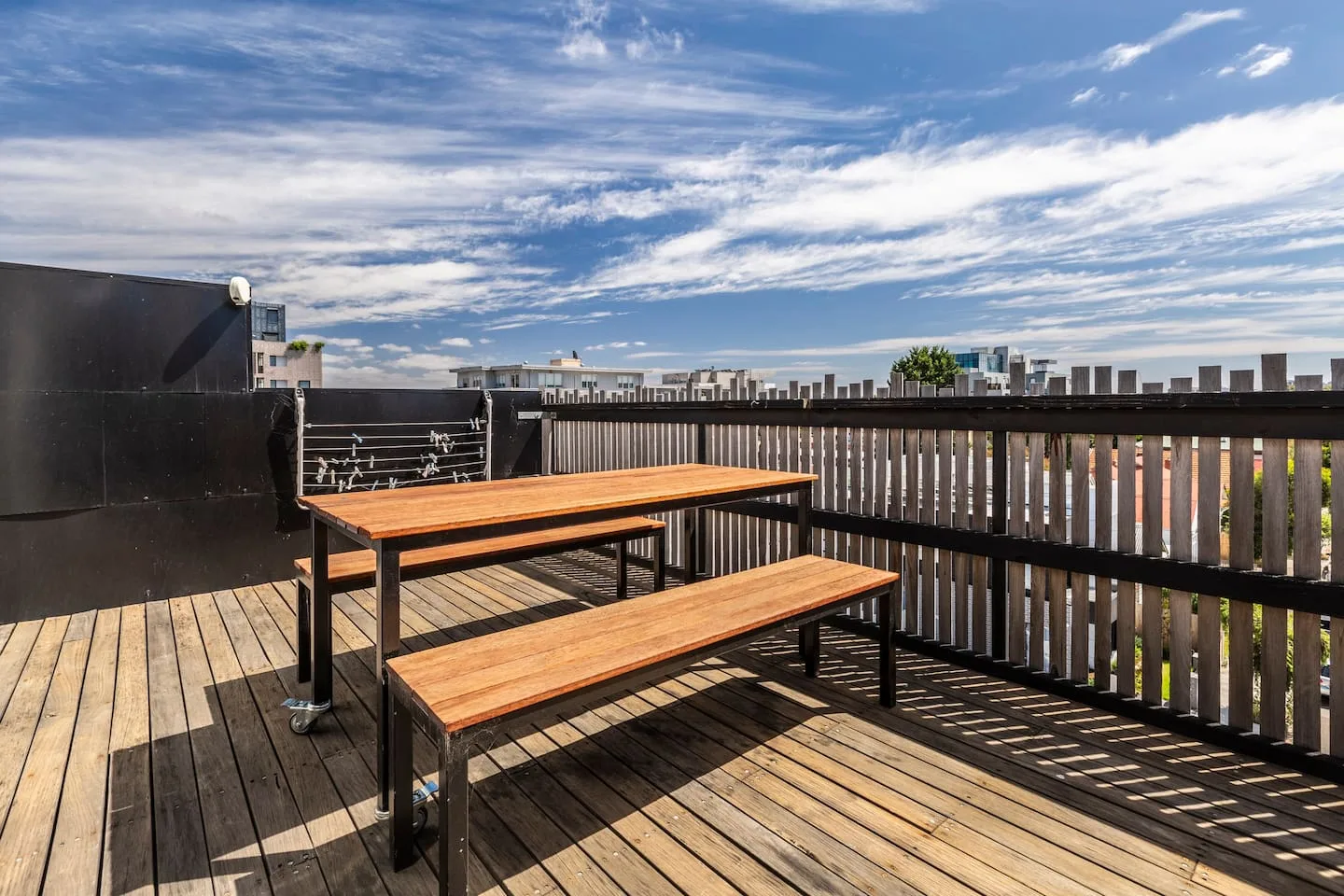 Una terraza de madera tiene una mesa de estilo pícnic y un banco junto a una barandilla oscura.