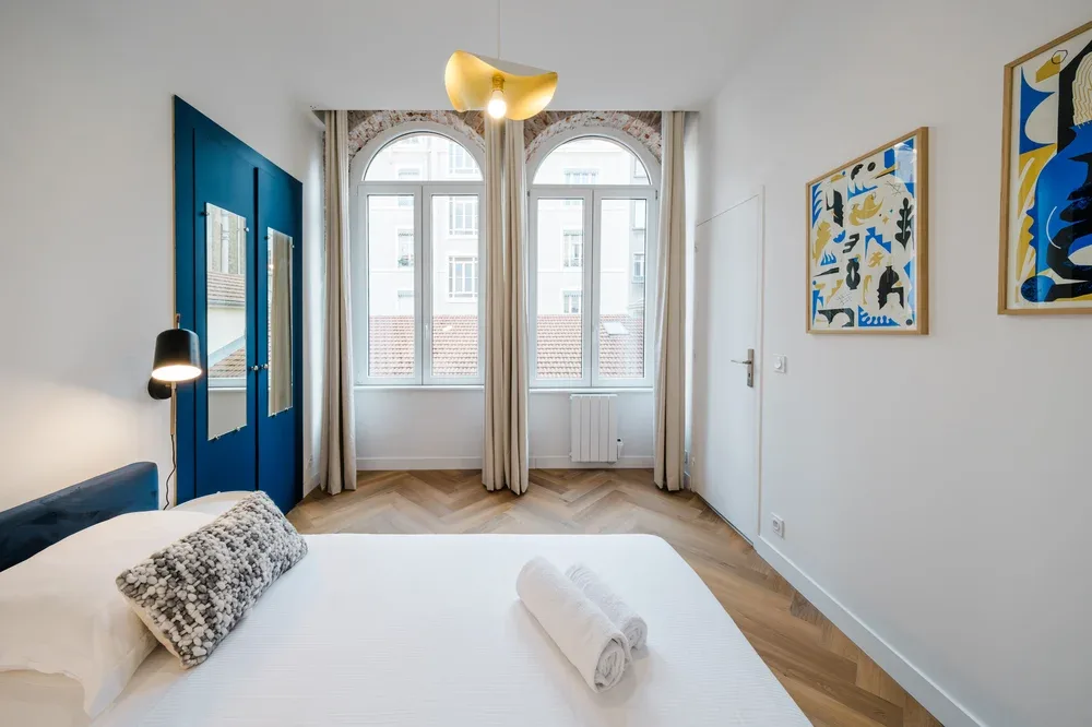 A bright bedroom with a neatly made bed and tall arched windows.