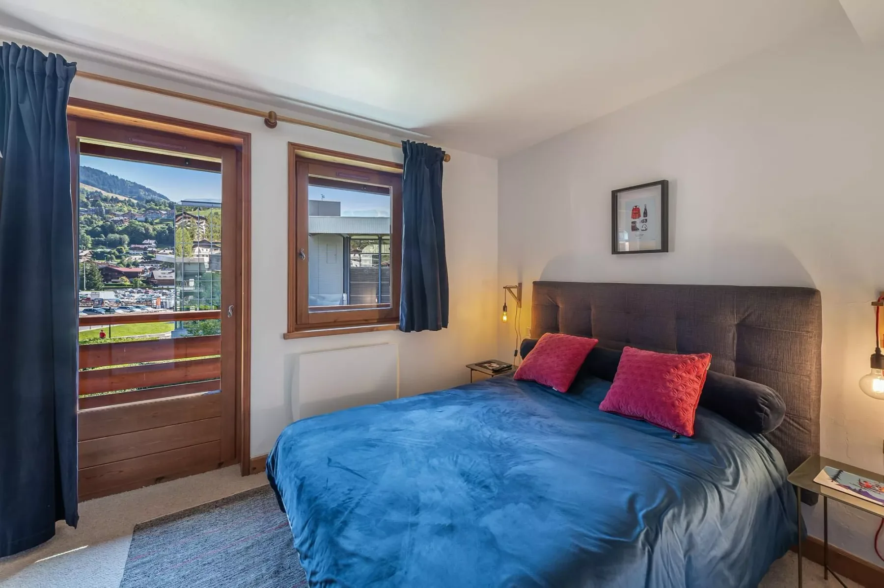A bedroom with a large bed, colorful pillows, and windows overlooking a hillside town.