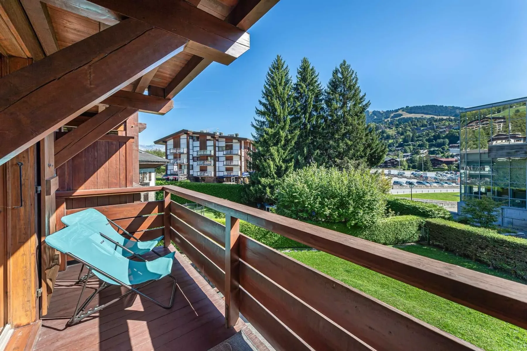 A wooden balcony overlooks greenery, nearby buildings, and distant hills.