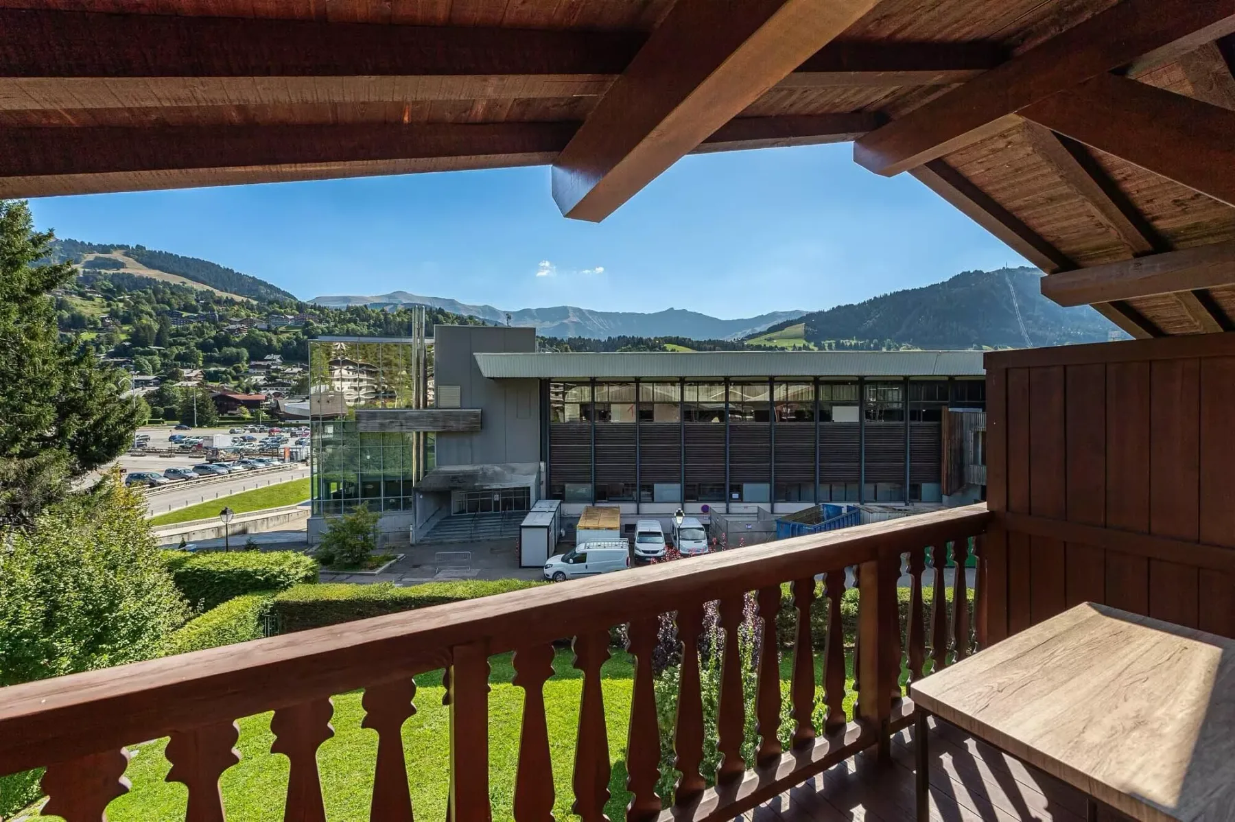 A wooden balcony overlooks a modern building and a green mountain landscape.