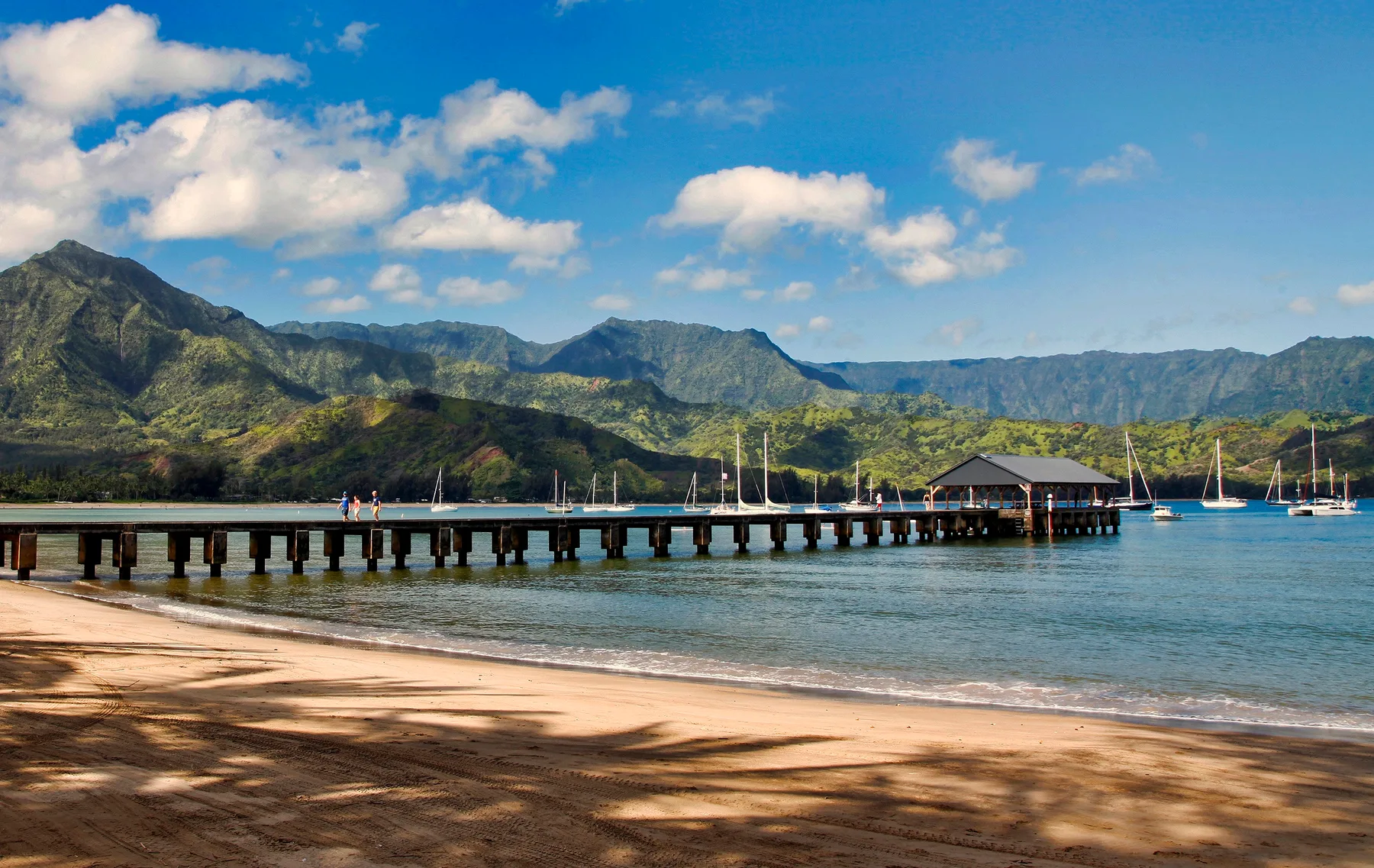 A sandy beach borders calm water, with a long pier and green mountains beyond.