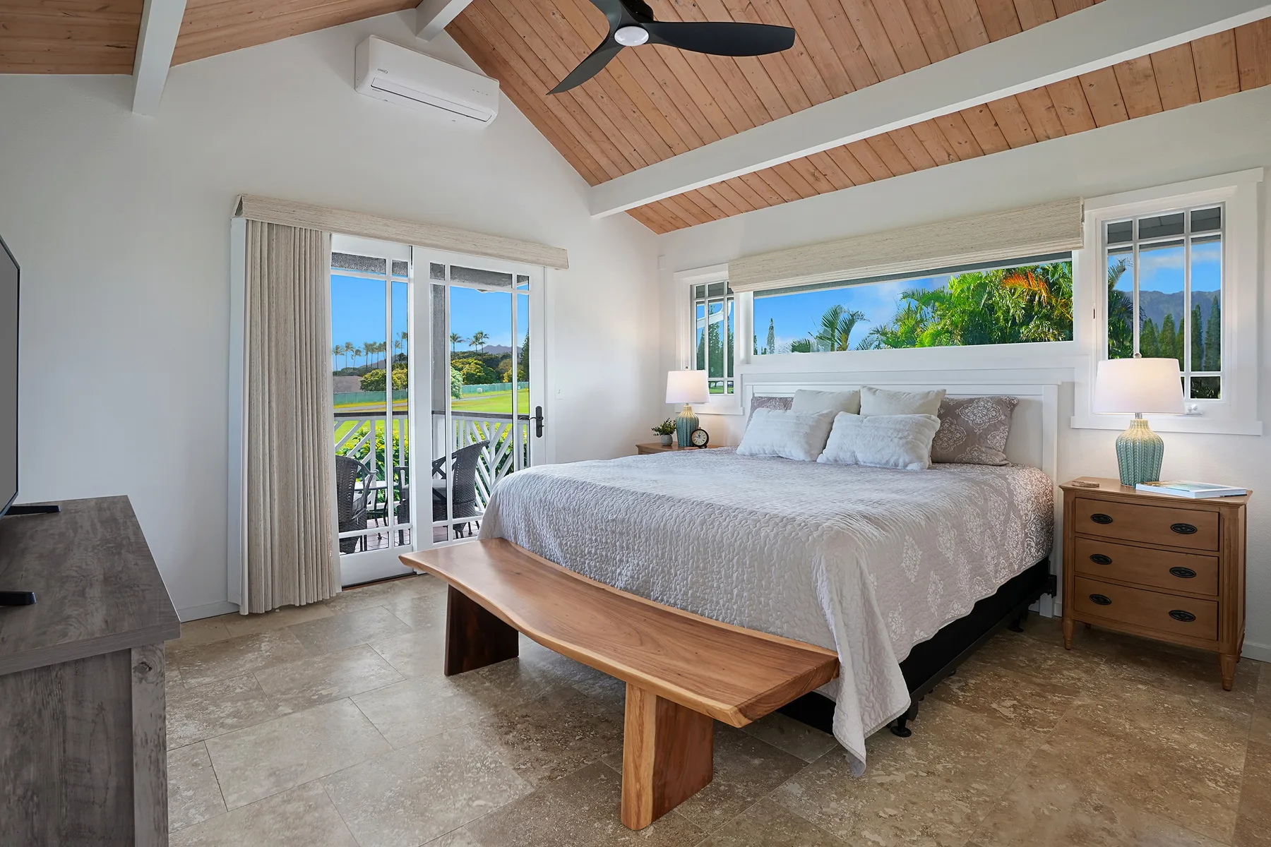 A bright bedroom with a large bed, wooden ceiling, and windows overlooking greenery.
