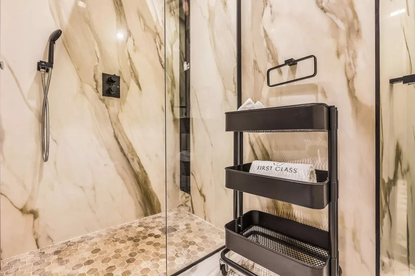 Un baño con acabado de mármol tiene una ducha de vidrio y un estante negro.