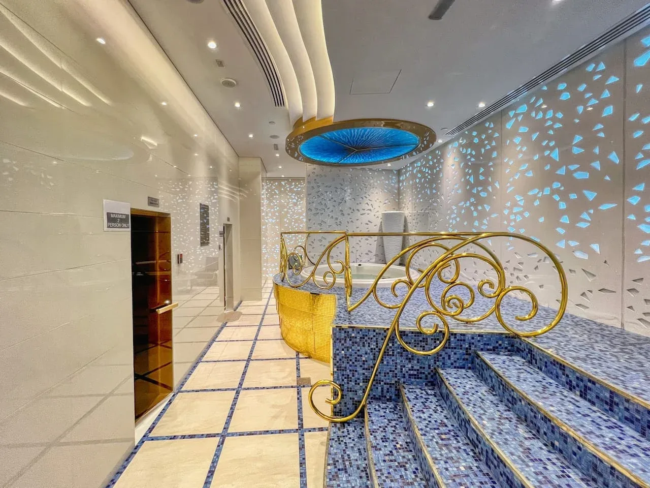 Zona de piscina interior con azulejos, una barandilla dorada decorativa y una luz azul en el techo.