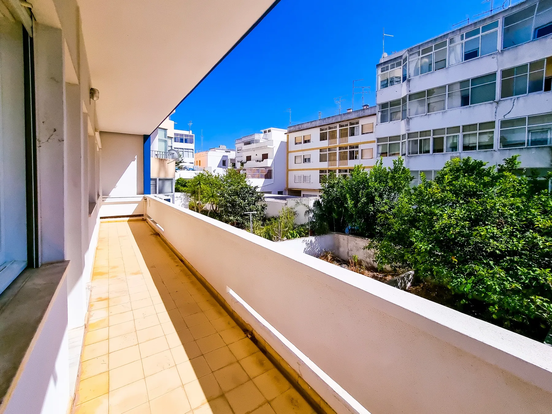A sunny balcony overlooks nearby buildings and leafy trees.