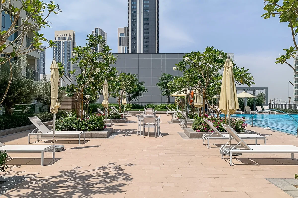 Una terraza con piscina tiene tumbonas, sombrillas cerradas y árboles.