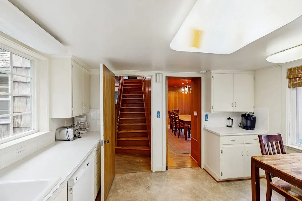 A bright kitchen with white cabinets and doorways leading to a staircase and dining area.