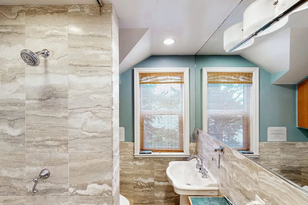 A bathroom with a tiled shower and a sink beneath two windows.