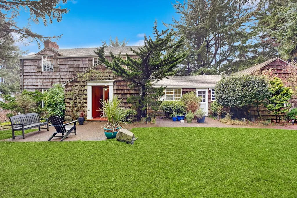 A garden lawn and patio sit in front of a shingled house surrounded by trees.