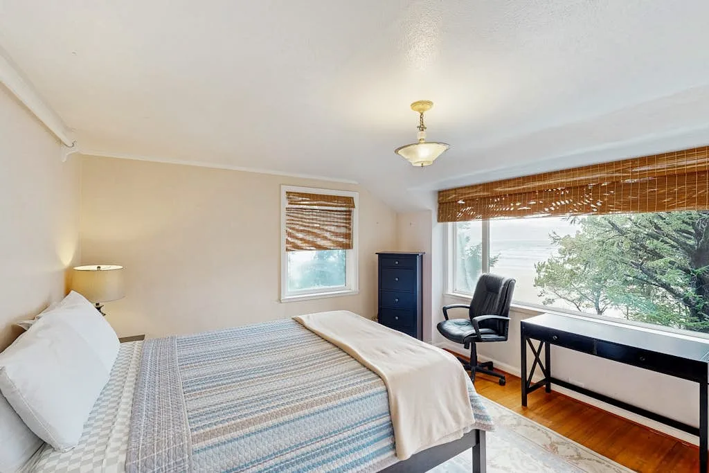 A bedroom with a bed and a desk beside a large window.