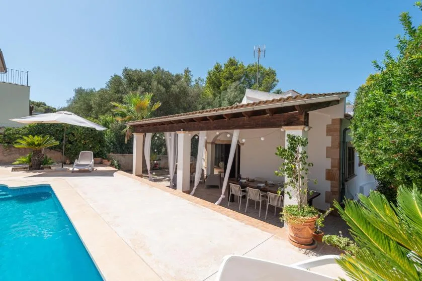 A poolside patio with a shaded dining area and surrounding greenery.