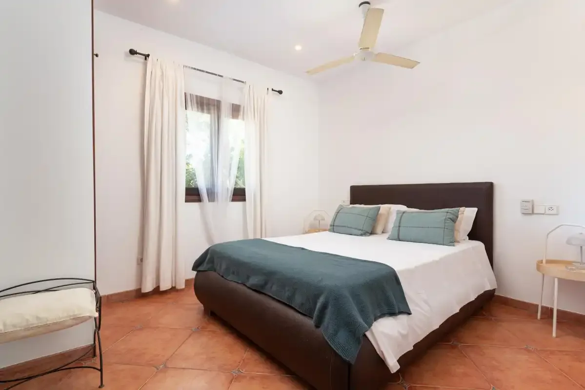 A bright bedroom with a double bed and a window with light curtains.