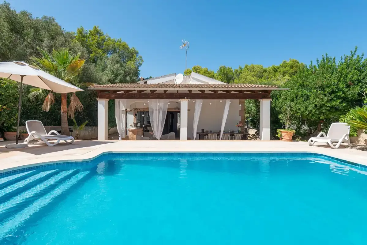 An outdoor swimming pool faces a covered patio surrounded by greenery.
