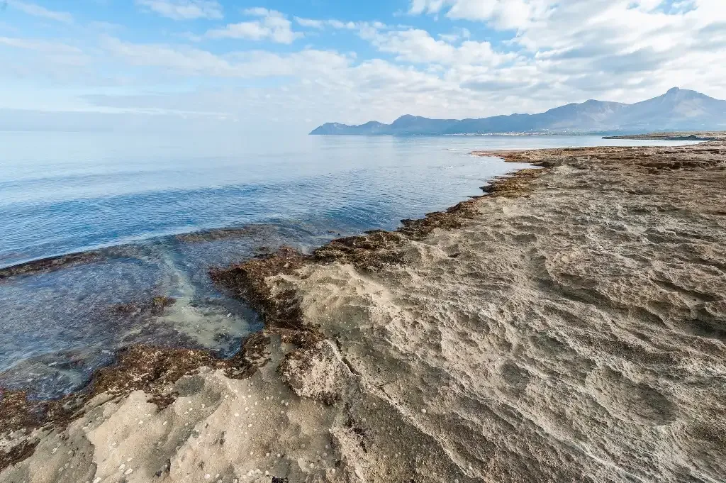 A rocky shoreline meets calm blue water, with distant mountains on the horizon.