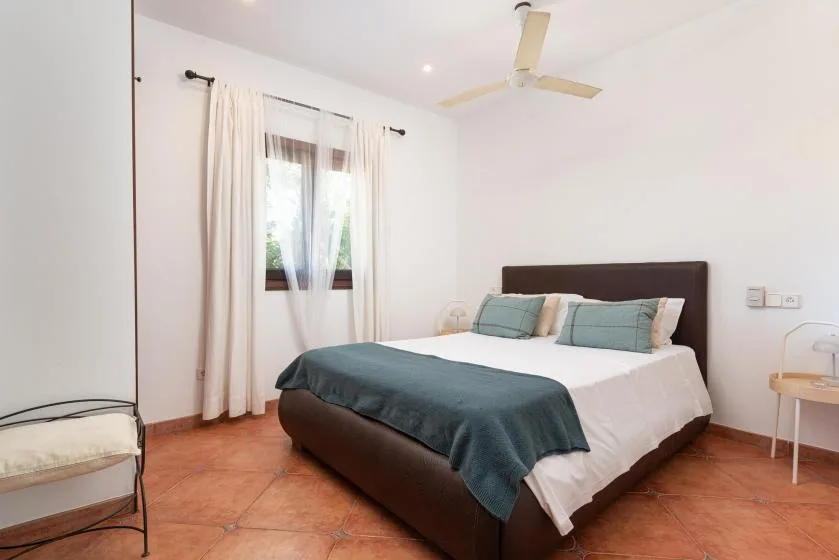 A bedroom with a bed dressed in white linens and a teal blanket beside a curtained window.