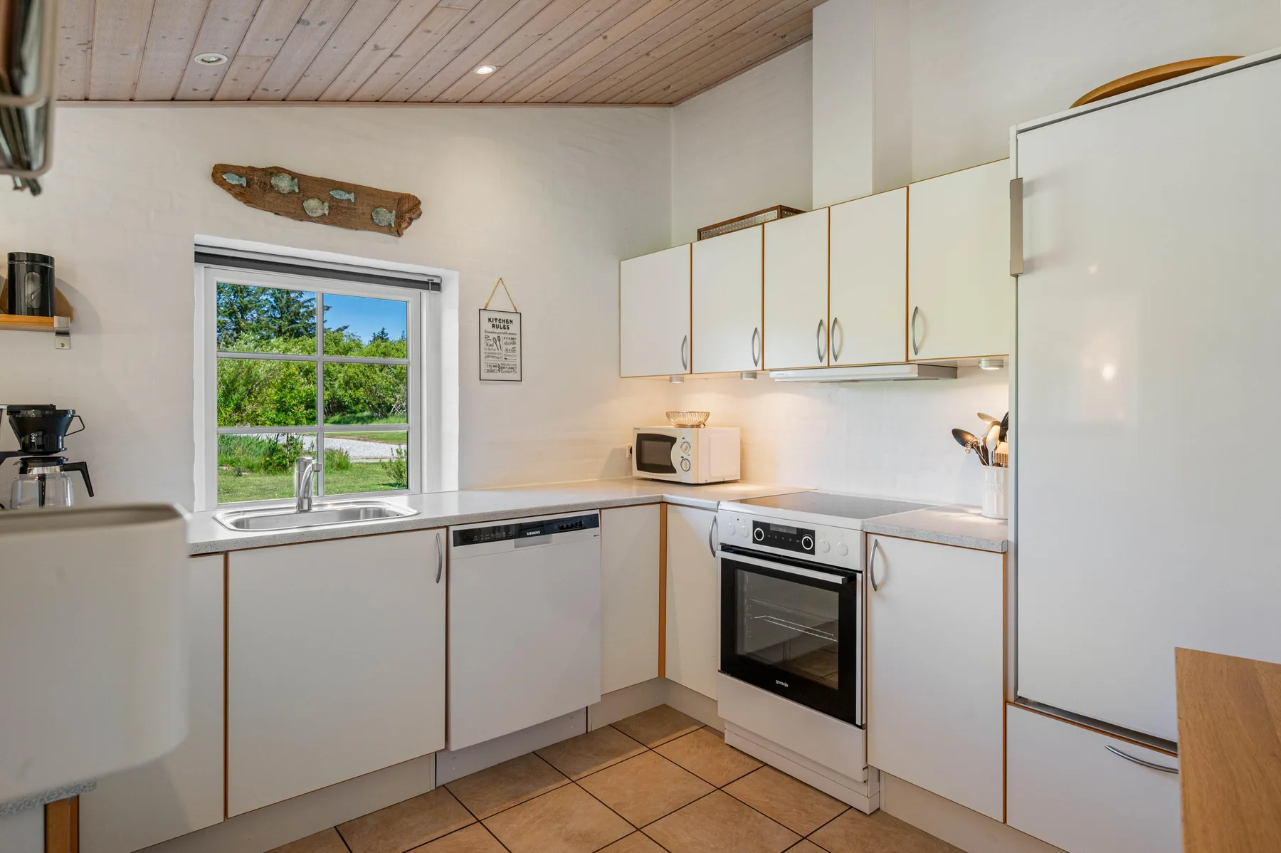 Una cocina luminosa con gabinetes blancos, suelo de baldosas y una ventana con vistas a la vegetación.