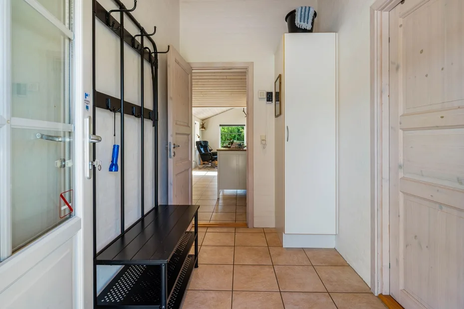 Un pasillo con suelo de baldosas, banco y percheros conduce a una habitación luminosa.