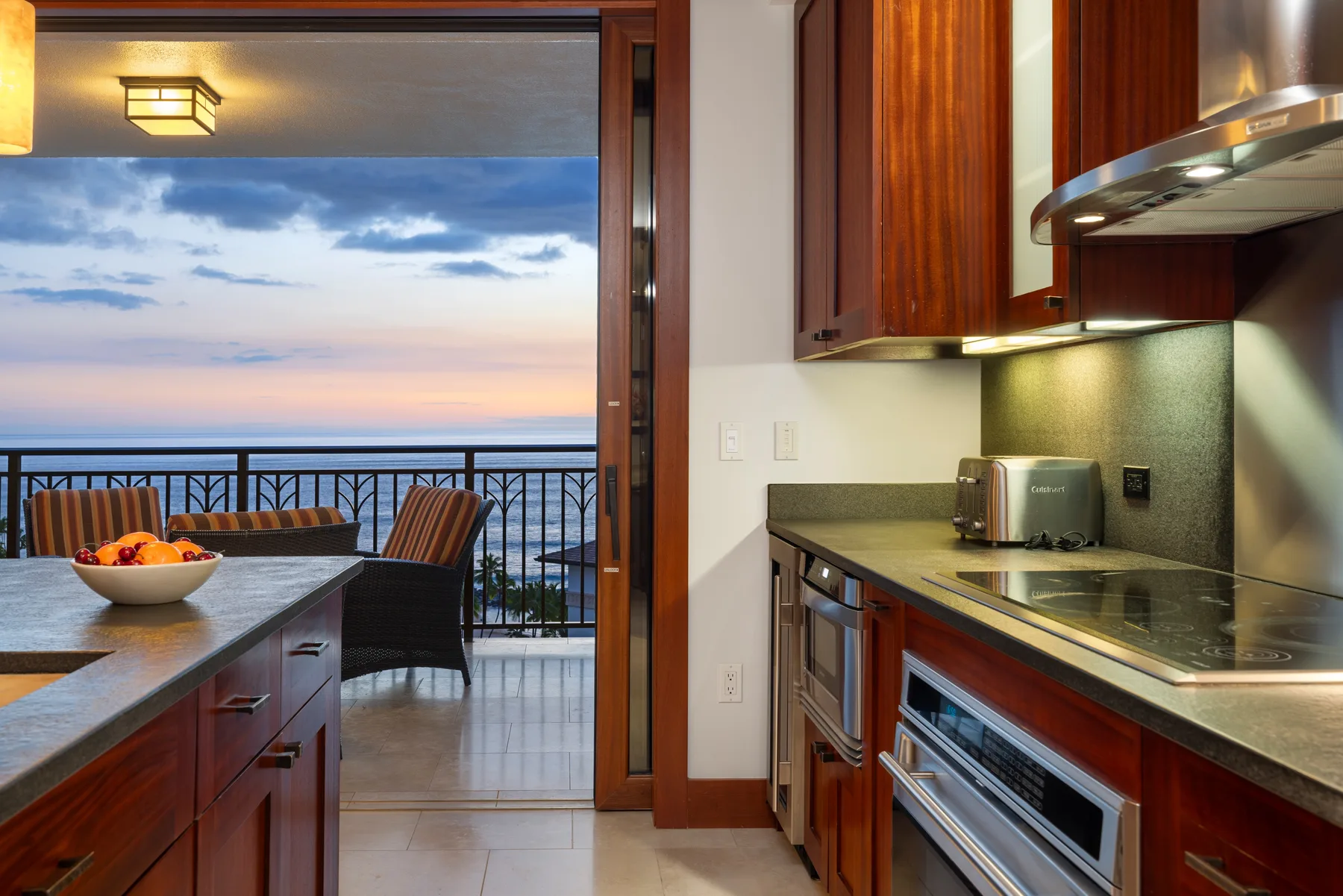 A kitchen opens onto a dining area with a balcony overlooking the water at sunset.