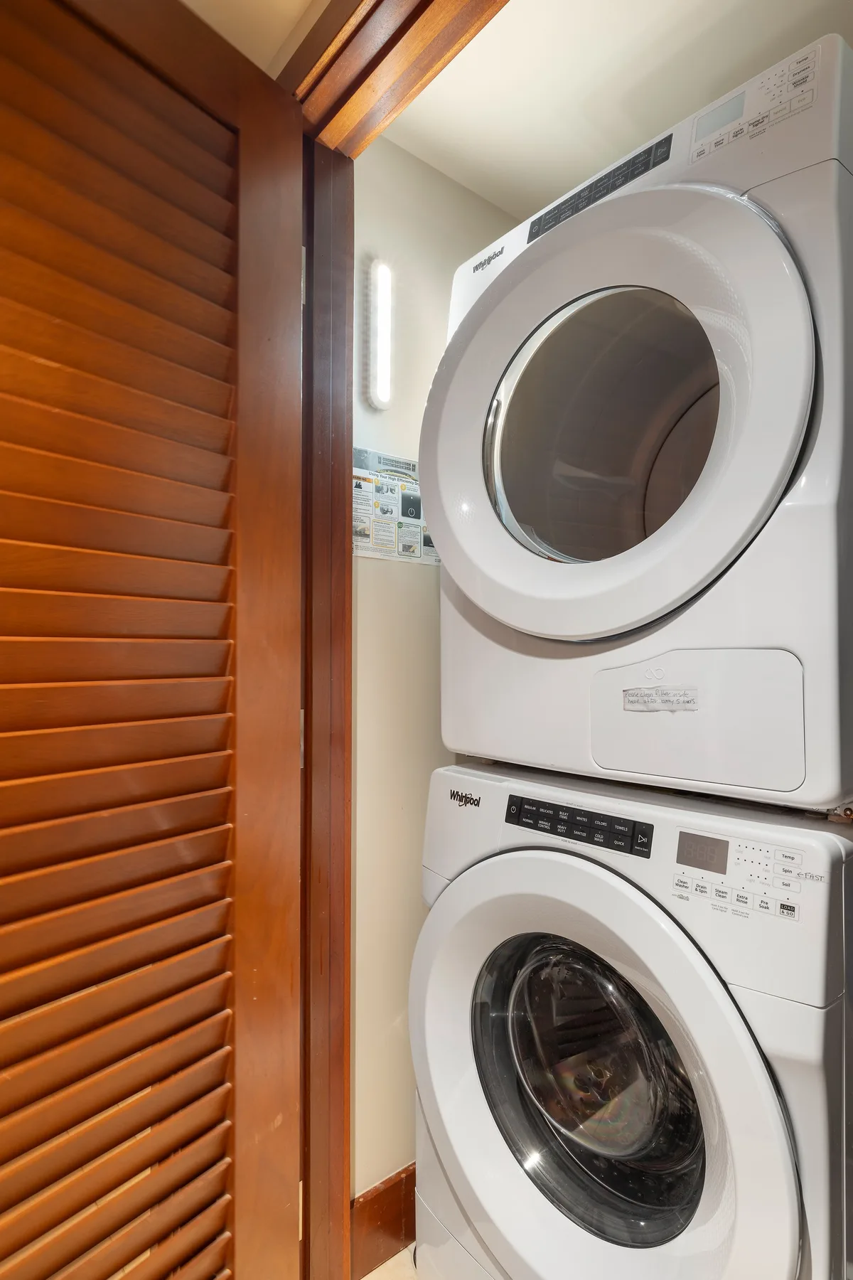 A laundry area with a stacked washer and dryer beside a wooden louvered door.